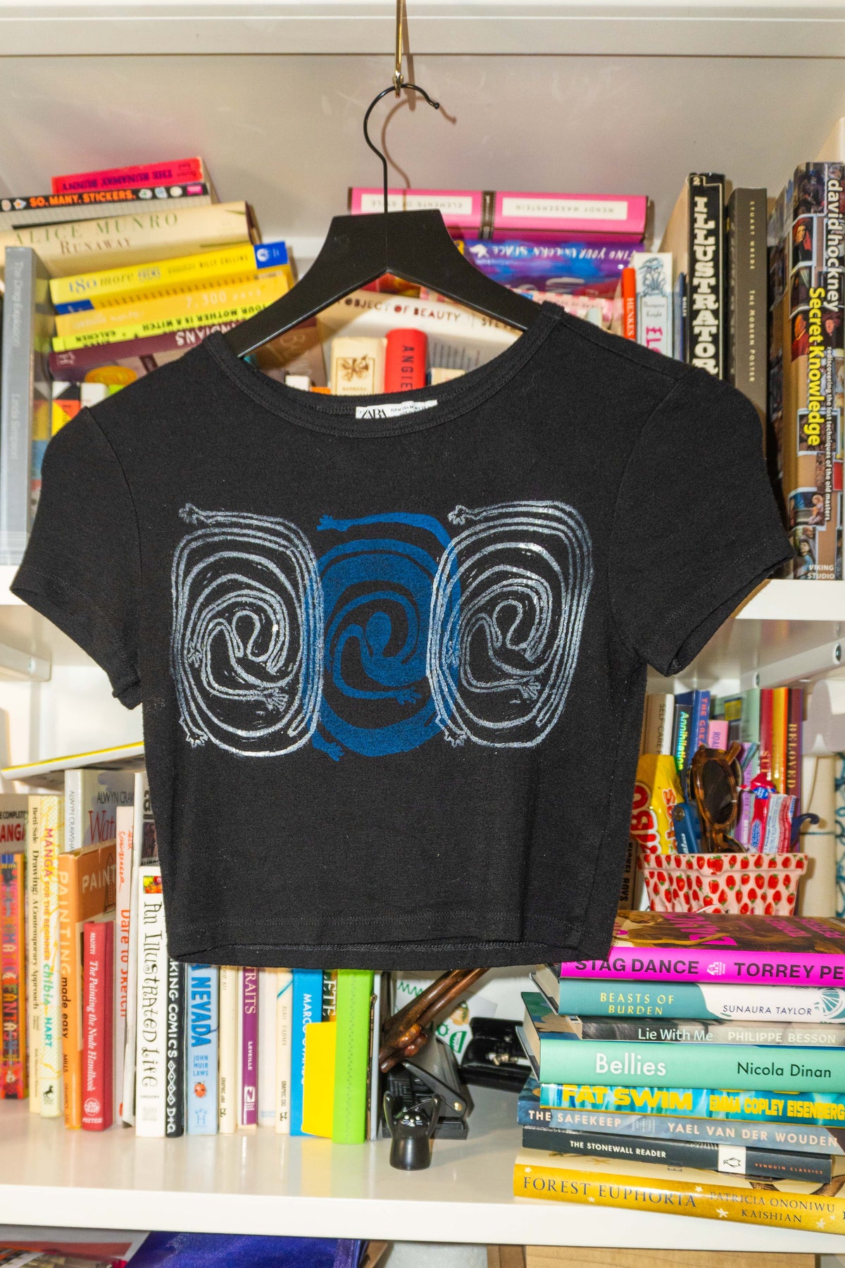 A black short-sleeve crop top t-shirt with a blue and white spiral linocut block print, hanging on a black hanger in front of a bookshelf.