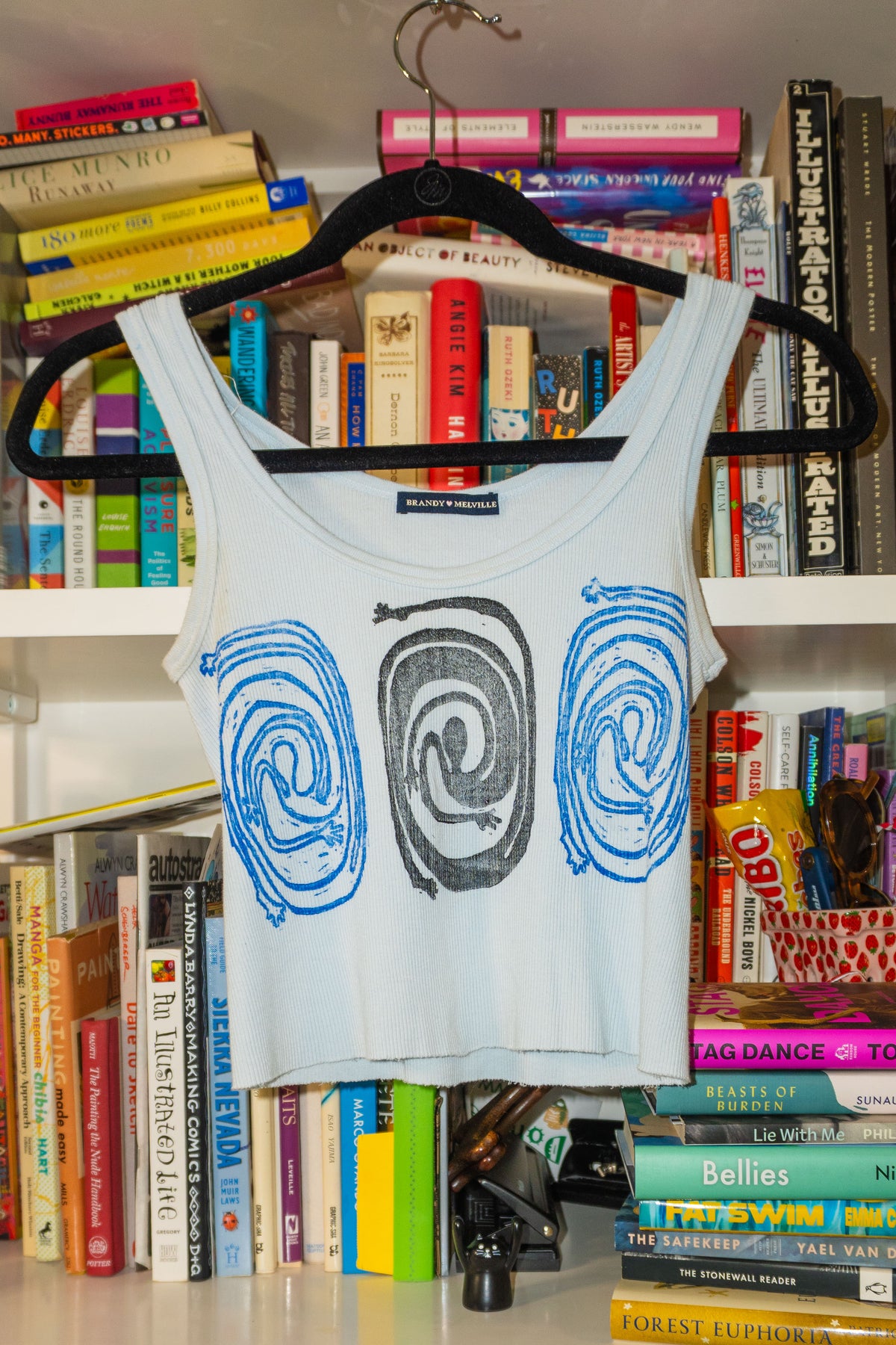 A light blue sleeveless crop top with a black and blue spiral linocut print, hanging on a black hanger in front of a bookshelf.