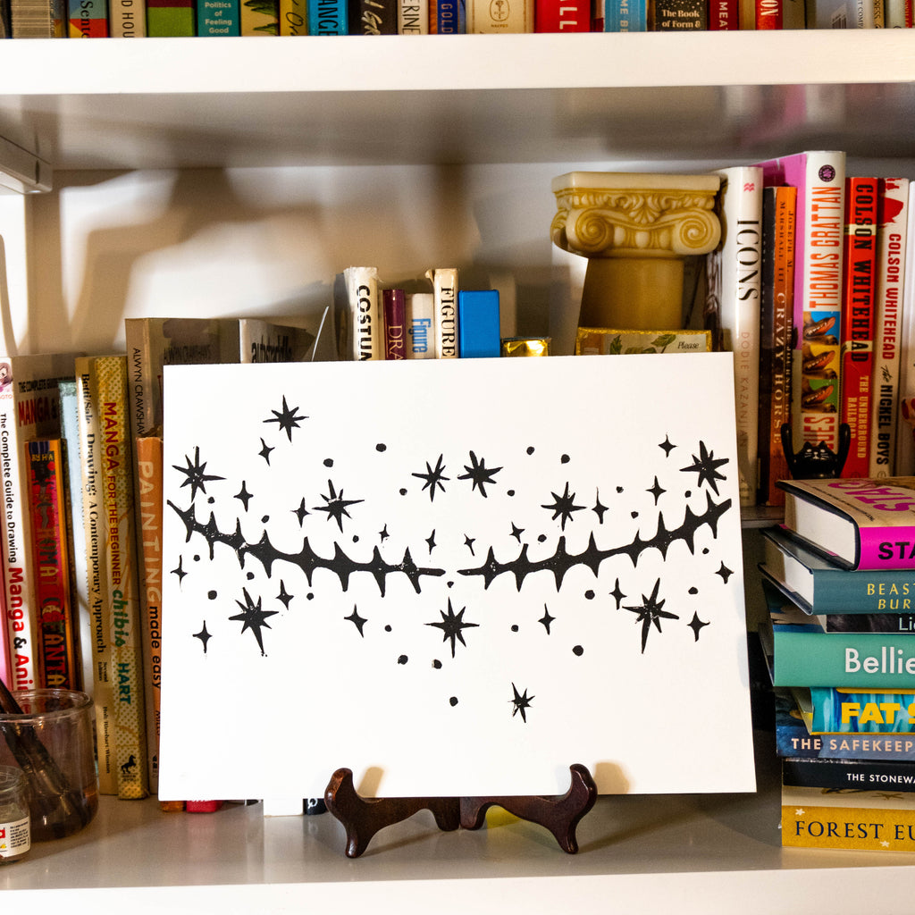 Starry Scars linocut print displayed on a small wooden easel in a bookshelf filled with knick knacks and books. 