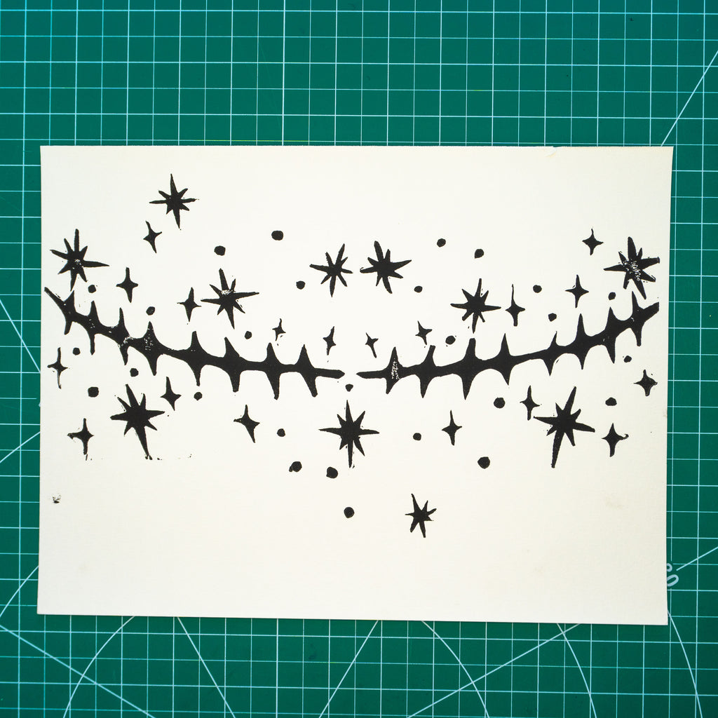 A white rectangular paper featuring a black in linocut design of top surgery scars with scattered star motifs around them, place on a green cutting mat with grid lines.