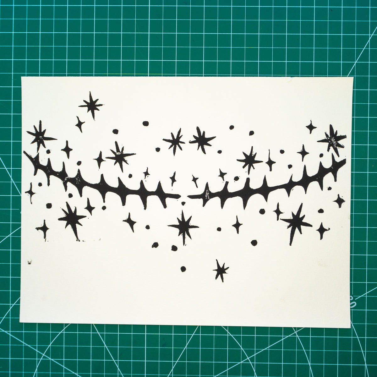 A white rectangular paper featuring a black in linocut design of top surgery scars with scattered star motifs around them, place on a green cutting mat with grid lines.