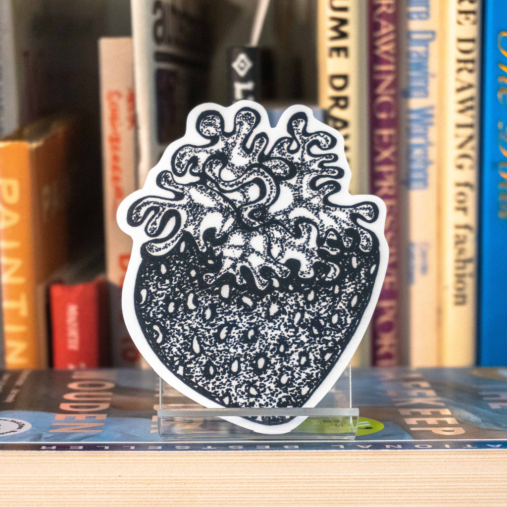 A large die-cut sticker featuring a grayscale tattoo-style strawberry displayed on a clear acrylic stand on top of a bookshelf with books in the background.