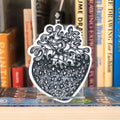 A large die-cut sticker featuring a grayscale tattoo-style strawberry displayed on a clear acrylic stand on top of a bookshelf with books in the background.