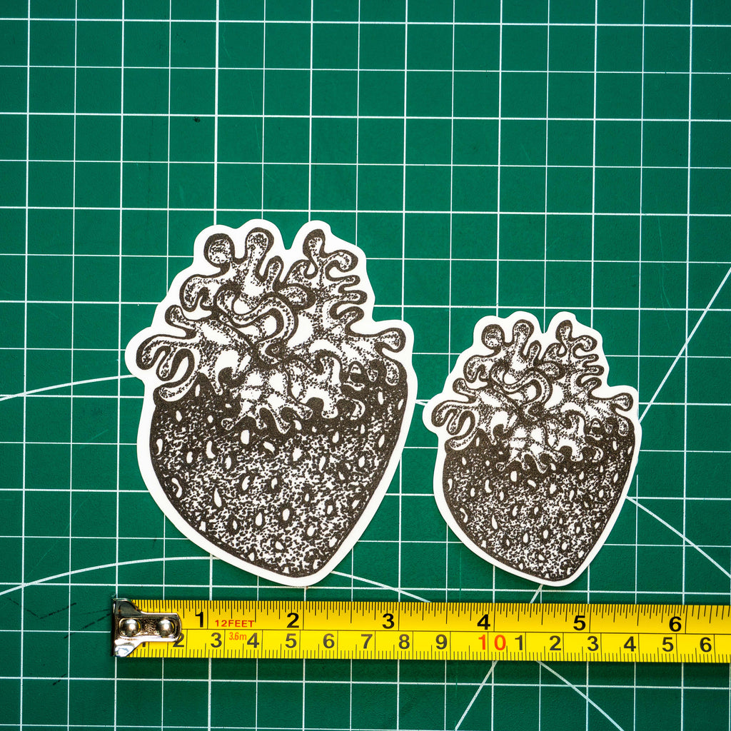 Two die-cut vinyl stickers of grayscale tattoo-style strawberries in different sizes, displayed on a green cutting mat with a yellow tape measure underneath for scale.