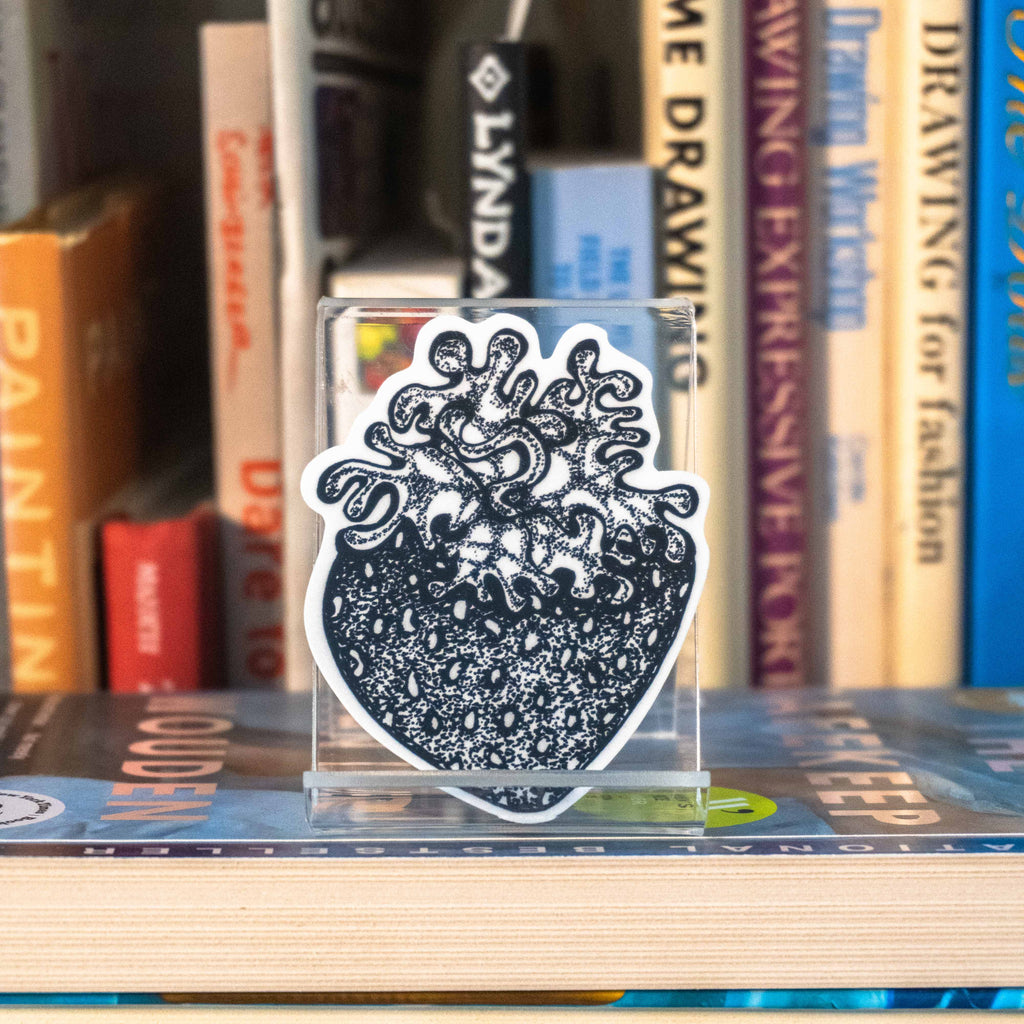 A small die-cut sticker featuring a grayscale tattoo-style strawberry displayed on a clear acrylic stand on top of a bookshelf with books in the background.