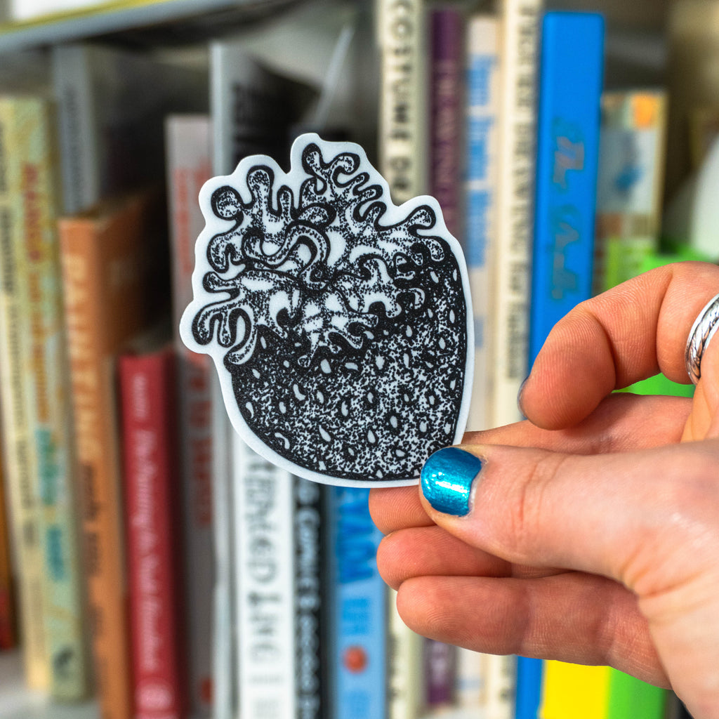 A hand with blue glitter nail polish holds a small die-cut sticker featuring a grayscale tattoo-style strawberry, with a blurred bookshelf in the background.