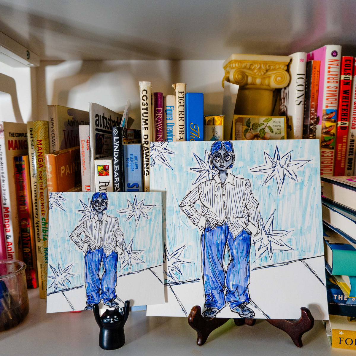 Two hand-drawn character art prints displayed on small stands on a shelf, with books and decorative items in the background.