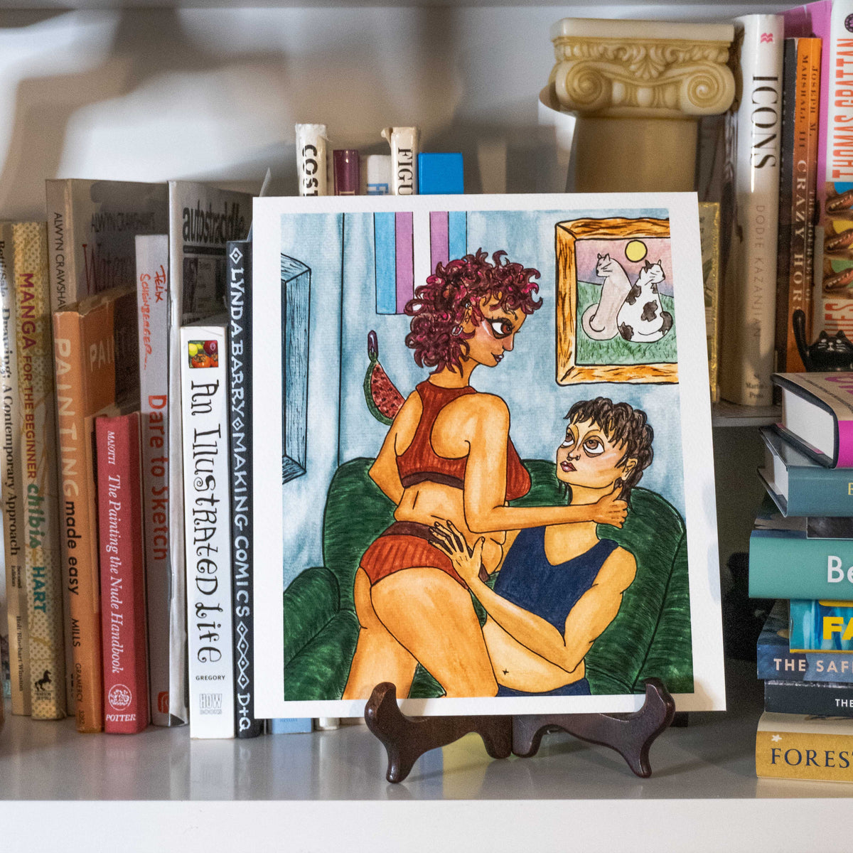 A square illustrated art print displayed on a small stand on a shelf, surrounded by books. The print features a colorful cartoon-style scene of two people sitting on a green couch, with one person wearing a red top and shorts and the other wearing a blue top.