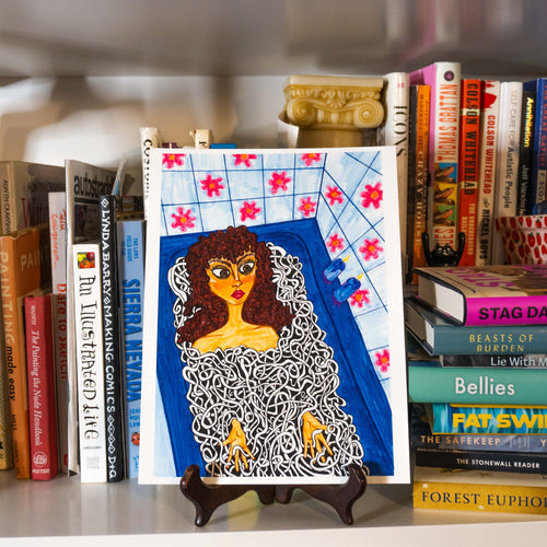 A colorful art print featuring a stylized illustration of a woman in a blue bathtub with pink flowers on the tiled wall, displayed on a small wooden easel stand on a bookshelf surrounded by books.