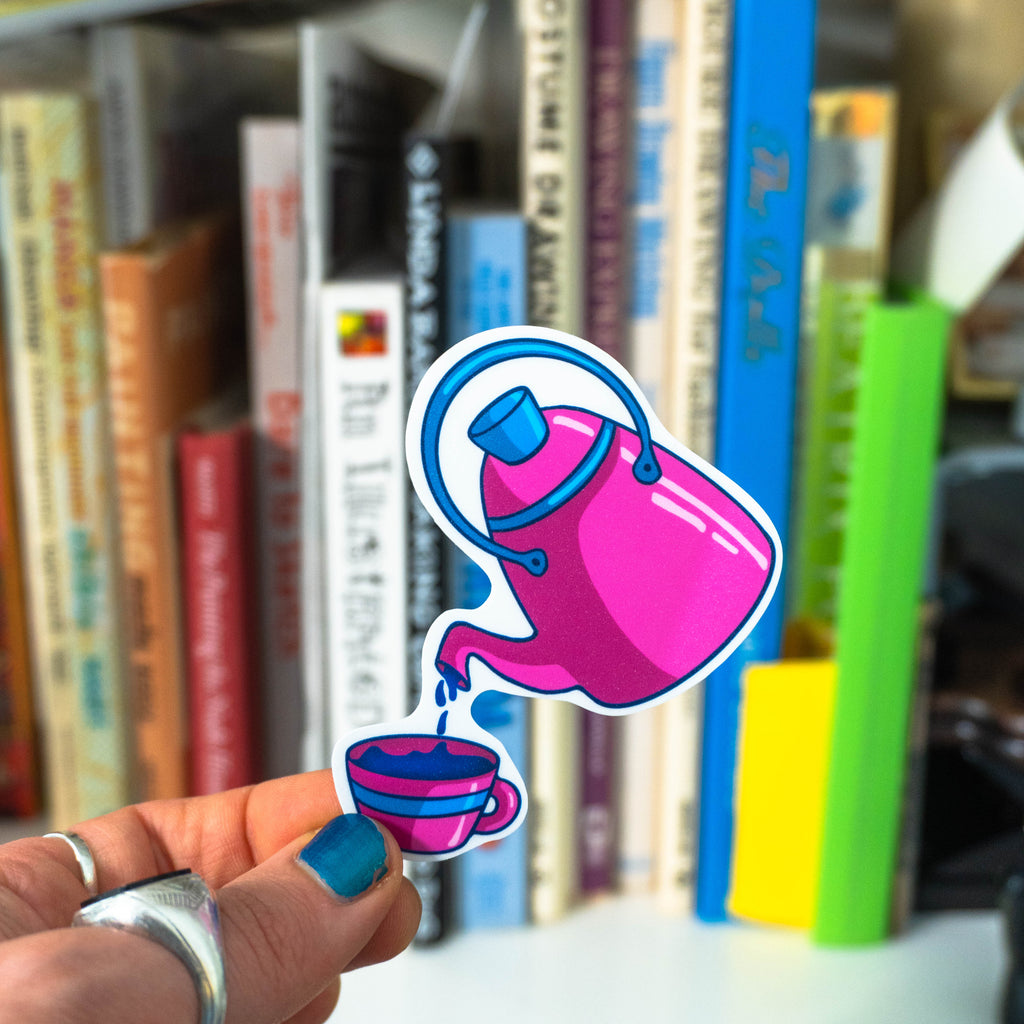 A hand with blue nail polish and silver rings holds a die-cut sticker featuring a pink teapot pouring into a blue and pink cup. The background is a blurred bookshelf with colorful books.