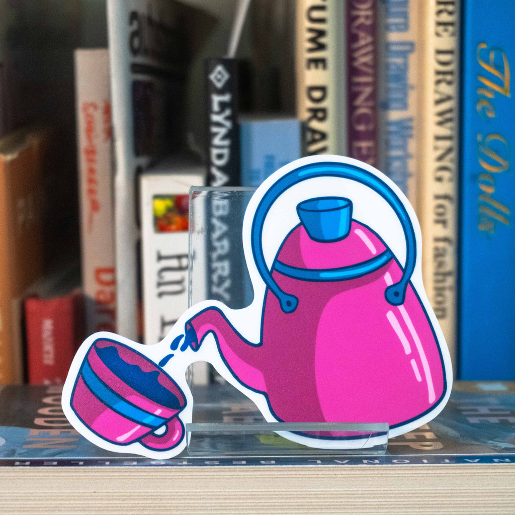 A large die-cut sticker featuring a pink teapot pouring into a matching cup, displayed on a clear acrylic stand on top of a bookshelf in front of blurred books.