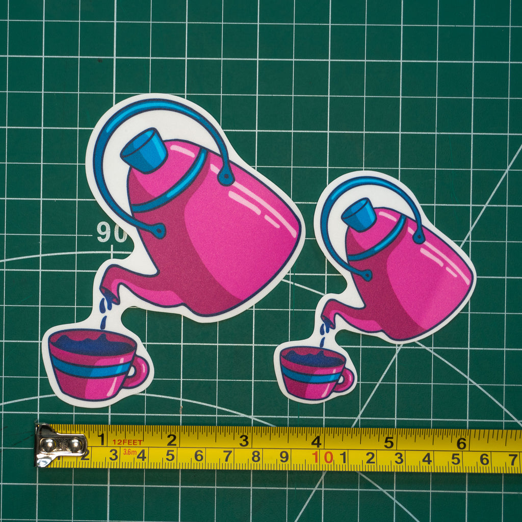 Two pink teapot and cup stickers in different sizes shown on a green cutting mat with a yellow tape measure at the bottom for scale.
