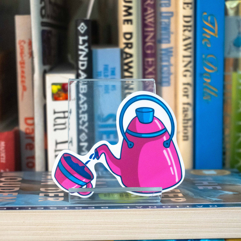 A small die-cut sticker featuring a pink teapot pouring into a matching cup, displayed on a clear acrylic stand on top of a bookshelf in front of blurred books.