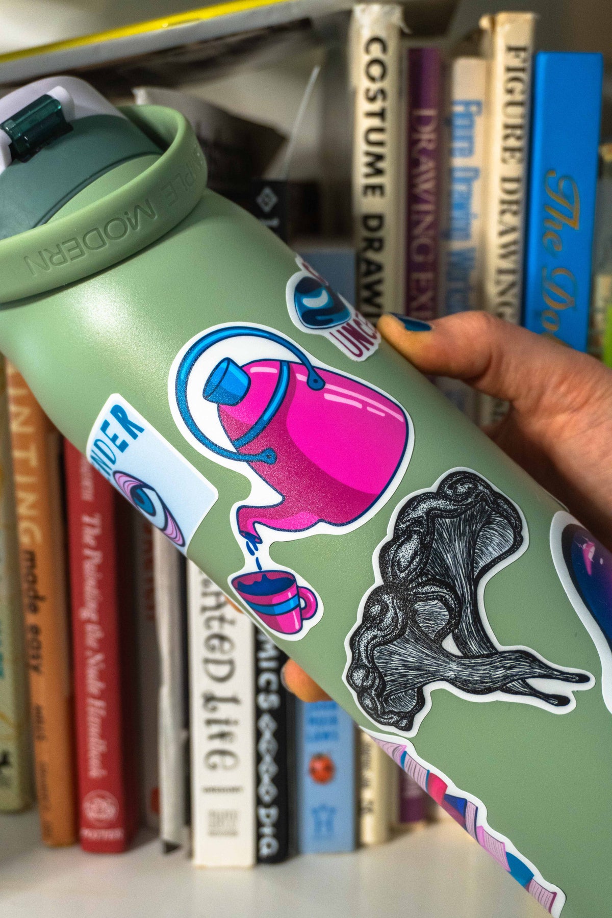 A sage green water bottle with colorful stickers, held by a hand in front of a bookshelf.