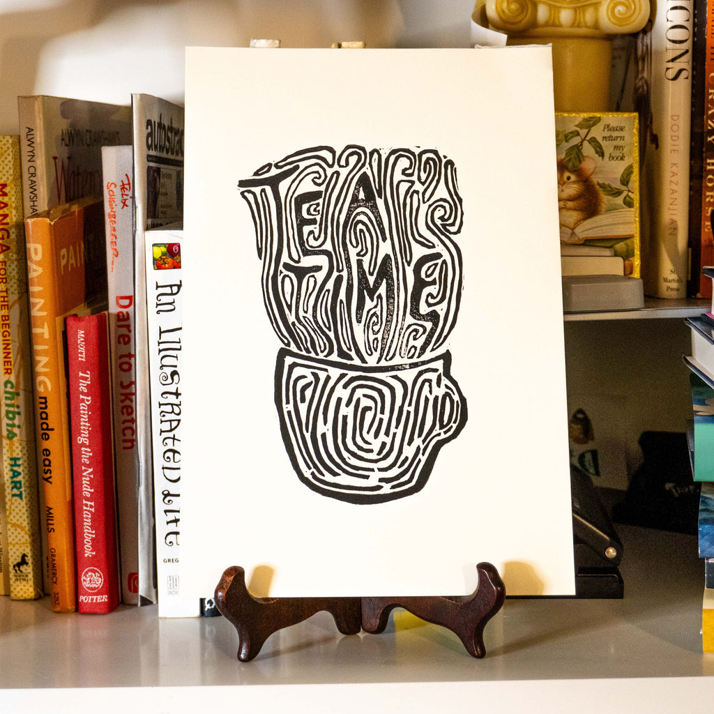 Tea Time Linocut print displayed on a small wooden easel stand on a shelf surrounded by books.