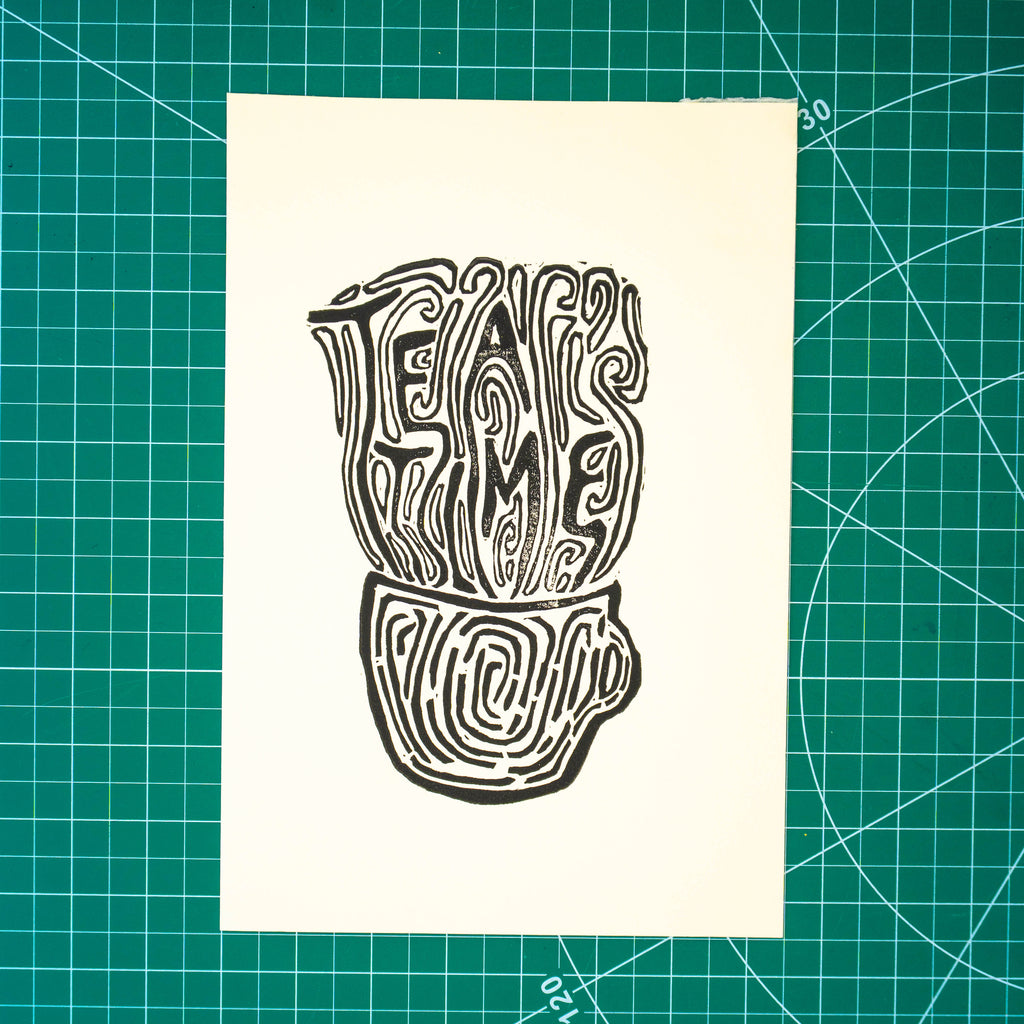 A square cream-colored art print featuring a black ink linocut illustration of a teapot and the words 'TEA TIME', displayed flat on a green cutting mat with grid lines.
