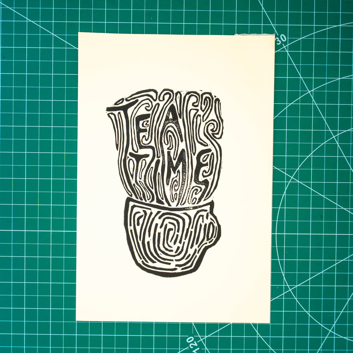 A square cream-colored art print featuring a black ink linocut illustration of a teapot and the words 'TEA TIME', displayed flat on a green cutting mat with grid lines.
