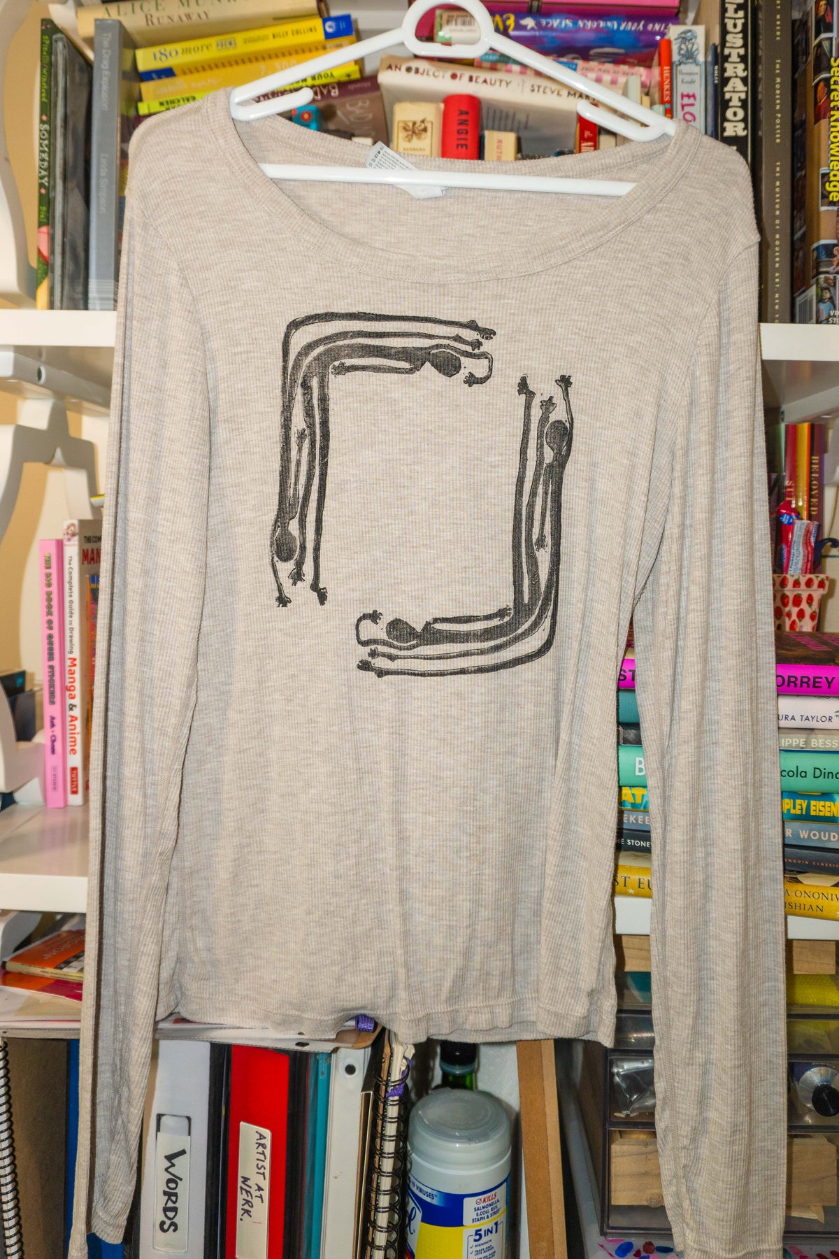 A light beige long-sleeve t-shirt with a black linocut print of a stylized square made of human figures, hanging on a white plastic hanger in front of a bookshelf.